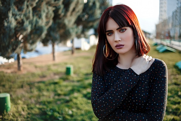 Hermosa joven con aspecto pesado y hosco en vestido negro fuera posando durante la puesta de sol