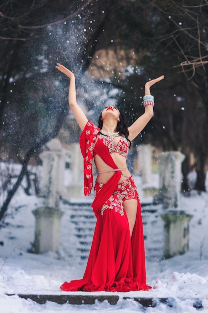 Hermosa joven bailando danza del vientre en vestido rojo en invierno en un parque en la nieve.