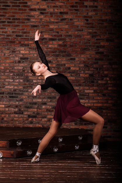 Hermosa joven bailarina de ballet clásico en pointe zapatos vistiendo un leotardo negro y falda en una pared de ladrillos