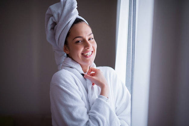 Hermosa joven en bata de baño y toalla en la cabeza de pie delante de la ventana y mirando a cámara.