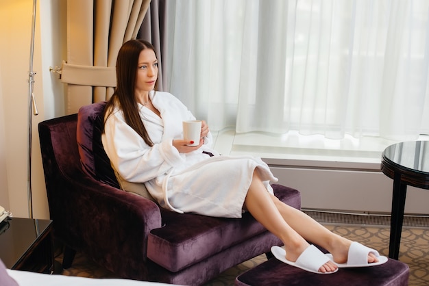 Foto una hermosa joven con una bata blanca bebe café en su habitación de hotel