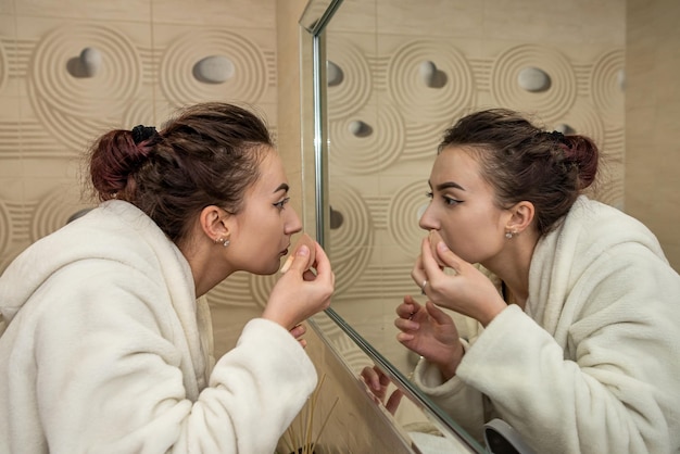 Hermosa joven con una bata blanca hace un maquillaje aburrido en un espejo espacioso El concepto de maquillaje en sí