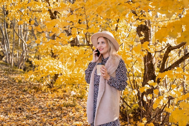 hermosa joven bebiendo café, hablando por teléfono en la naturaleza junto al lago en el parque de otoño en otoño