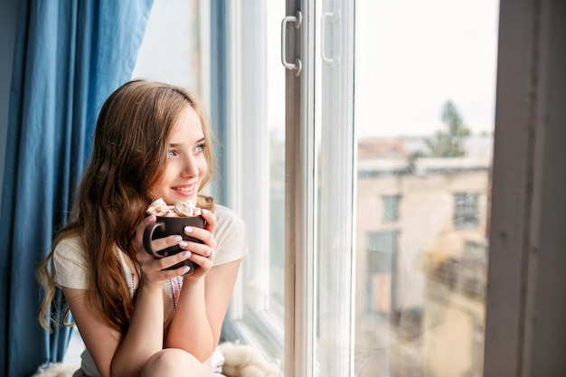 Hermosa joven bebiendo café y mirando por la ventana mientras está sentada en el alféizar de la ventana en casax9