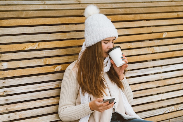 Hermosa joven bebiendo café, té de taza de plástico en invierno y hablando por teléfono móvil