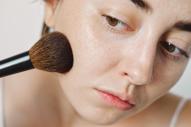 Una hermosa joven blanca con un pincel de maquillaje se maquilla en la cara. polvos faciales