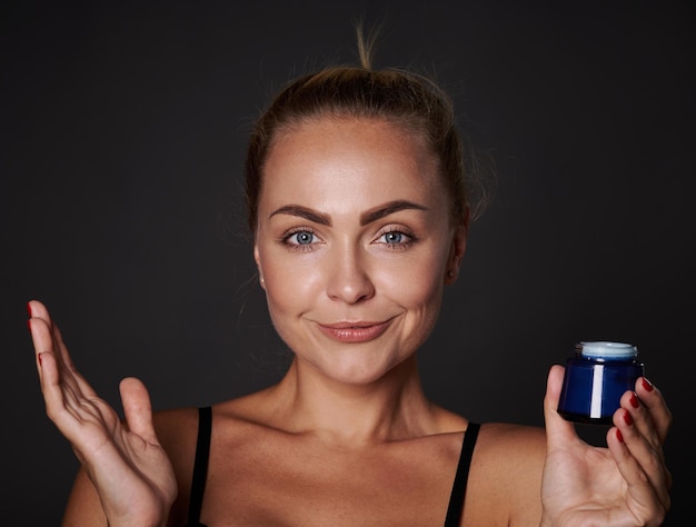 Hermosa joven de buen aspecto con una piel clara y fresca sonríe mirando a la cámara y sosteniendo una botella con crema facial hidratante antienvejecimiento aislada sobre fondo negro con espacio para copiar