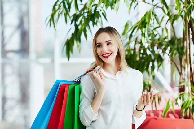 Hermosa joven con cabello castaño claro y labios rojos con blusa blanca y de pie con coloridas bolsas de compras, sosteniendo el teléfono móvil, concepto de compras, espacio de copia.