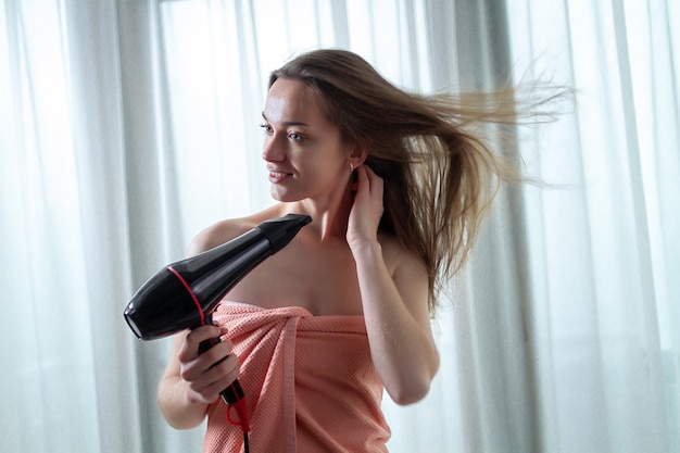 Hermosa joven de cabello castaño en una toalla de baño está usando un secador de pelo para secar su cabello largo después de la ducha en la habitación. Cuidado del cabello.