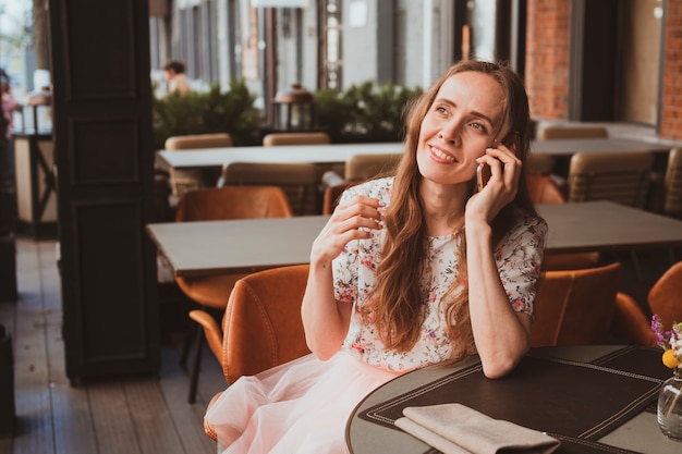 Hermosa joven con cabello largo está sentada en un café y hablando por el teléfono inteligente