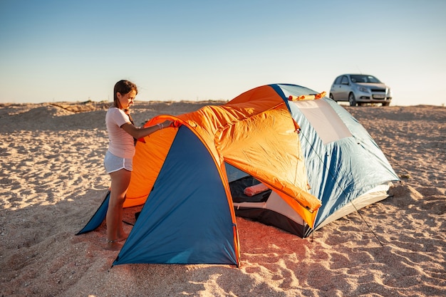 Hermosa joven con cabello oscuro instala una carpa en la playa. Autocaravana en una playa desierta