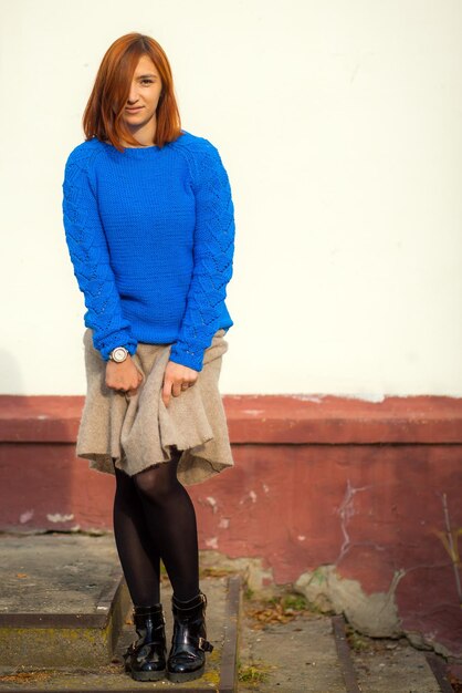 Una hermosa joven de cabello rojo con un suéter azul hecho de lana natural y falda beige de lana sonriendo y posando en una calle de la ciudad en el día de otoño