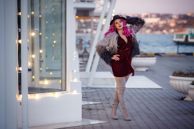 Una hermosa joven con cabello rosado y un abrigo inusual camina por la ciudad nocturna disfrutando de la luz de las guirnaldas y la luz del atardecer