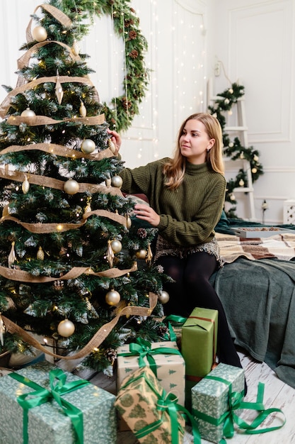 Una hermosa joven con cabello rubio se sienta cerca de un árbol de Navidad y decora con juguetes el Año Nuevo es pronto un ambiente navideño en un hogar acogedor