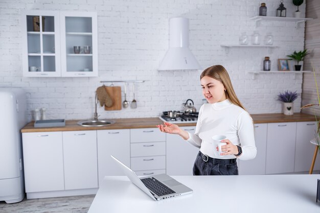 Una hermosa joven de cabello rubio con un suéter de cuello alto blanco se encuentra en una cocina blanca y trabaja en una computadora portátil, toma café. Trabaja de forma remota desde casa