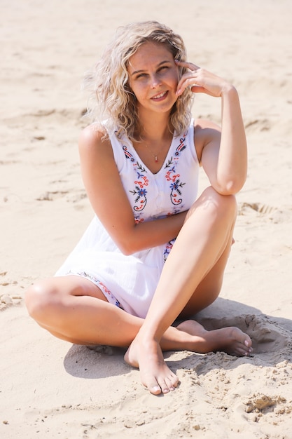 Hermosa joven con cabello rubio con un vestido de verano se sienta en la arena de la orilla