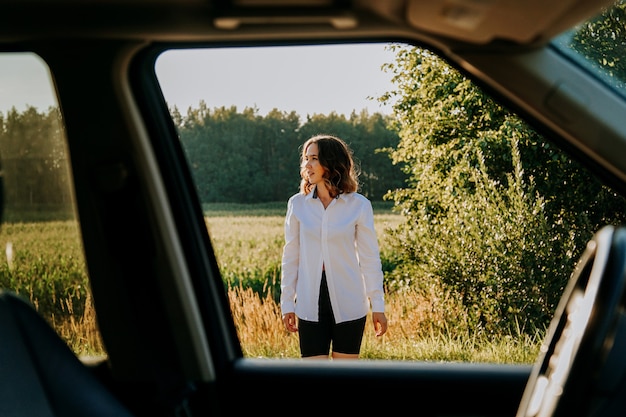 Una hermosa joven con una camisa blanca descansa fuera de la ciudad. Al aire libre cerca del bosque y el campo. Foto a través de la ventanilla del coche. Viaje en coche