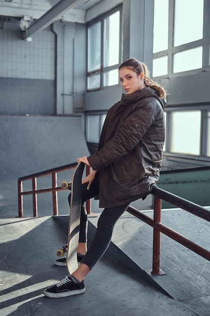 Una hermosa joven con una camisa a cuadros y un abrigo con su patineta sentada en un riel de molienda en el parque de patinaje en el interior.