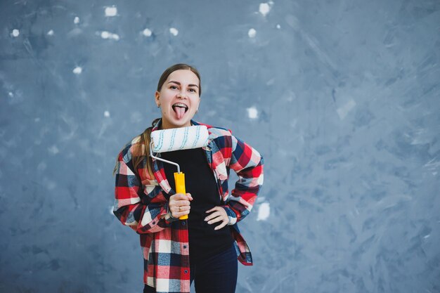 Una hermosa joven con una camisa a cuadros está renovando su habitación con un rodillo de pintura