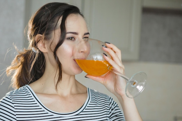 Una hermosa joven con una camiseta a rayas bebe rodajas de naranja y jugo en la cocina de un vaso