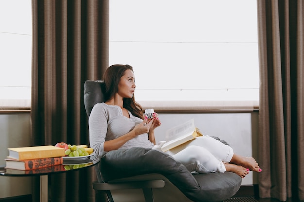 La hermosa joven en casa sentada en una silla moderna frente a la ventana, relajándose en su sala de estar, leyendo un libro y tomando café o té