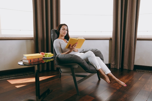 La hermosa joven en casa sentada en una silla moderna frente a la ventana, relajándose en su sala de estar y leyendo un libro