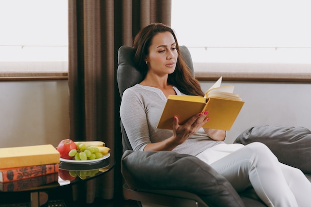 La hermosa joven en casa sentada en una silla moderna frente a la ventana, relajándose en su sala de estar y leyendo un libro