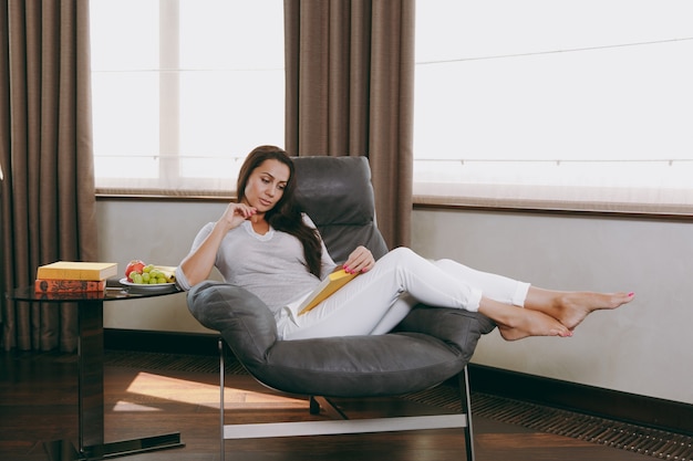 Foto la hermosa joven en casa sentada en una silla moderna frente a la ventana, relajándose en su sala de estar y leyendo un libro