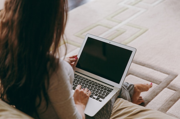 La hermosa joven en casa sentada en el sofá, relajándose en su sala de estar y trabajando con un portátil