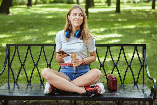Hermosa joven caucásica de unos 25 años con cabello rubio y auriculares está sentada en un banco en el parque público.