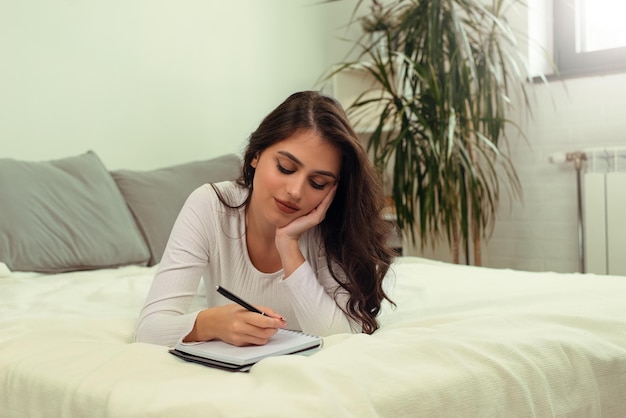 Hermosa joven caucásica acostada en la cama y escribiendo un diario