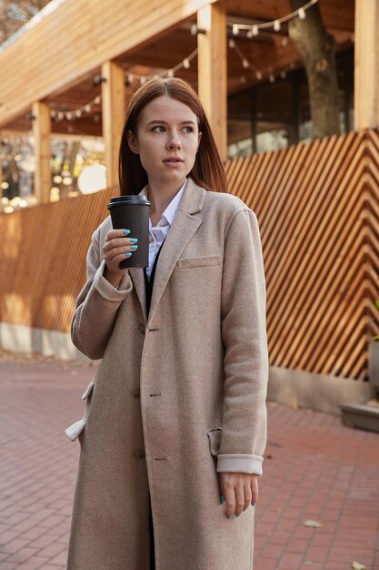 Hermosa joven caucásica en elegante abrigo beige tomando café al aire libre