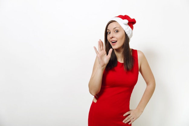 La hermosa joven caucásica feliz con vestido rojo y sombrero de Navidad que muestra el gesto de la mano y cuenta secretos sobre fondo blanco. Retrato aislado de la muchacha de Santa. Concepto de vacaciones de año nuevo 2018