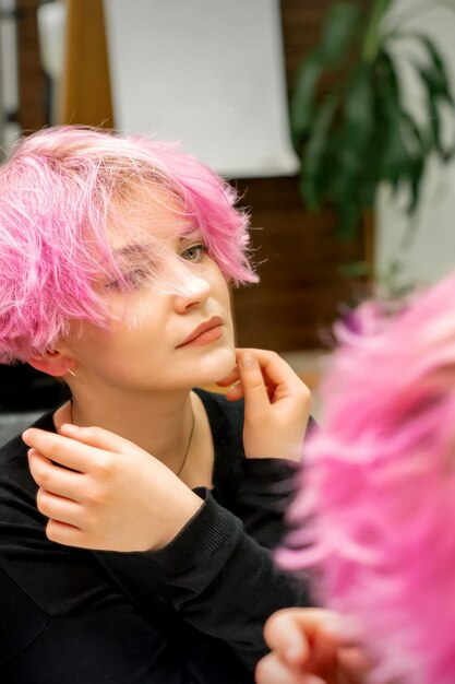 La hermosa joven caucásica con un nuevo peinado rosa corto mirando su reflejo en el espejo revisando el peinado en un salón de peluquería