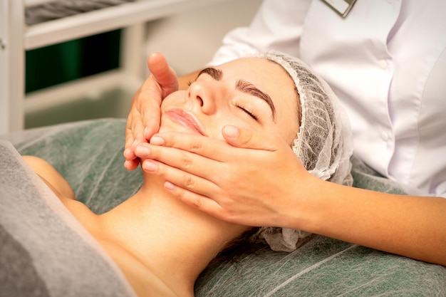 Hermosa joven caucásica recibiendo un masaje facial con los ojos cerrados en el salón de spa de cerca Concepto de tratamiento relajante