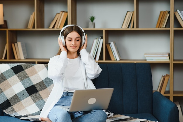 Foto hermosa joven caucásica en ropa casual disfrutando de la música y sonriendo mientras descansa en casa mujer joven con auriculares usando una computadora portátil en casa