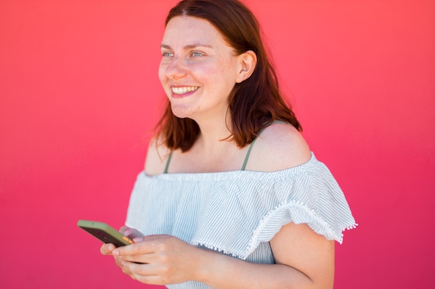 Hermosa joven caucásica sonriendo y usando el teléfono inteligente en la pared rosa con espacio de copia