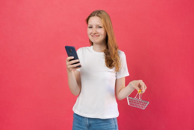 Hermosa joven caucásica sosteniendo un pequeño carrito de compras y un teléfono móvil