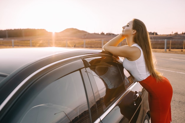 Hermosa joven cerca de un coche por la noche bajo el sol del atardecer en un estacionamiento vacío