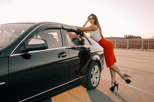 Hermosa joven cerca de un coche por la noche bajo el sol del atardecer