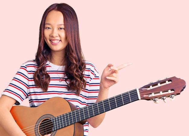 Hermosa joven china tocando la guitarra clásica sonriendo feliz señalando con la mano y el dedo a un lado