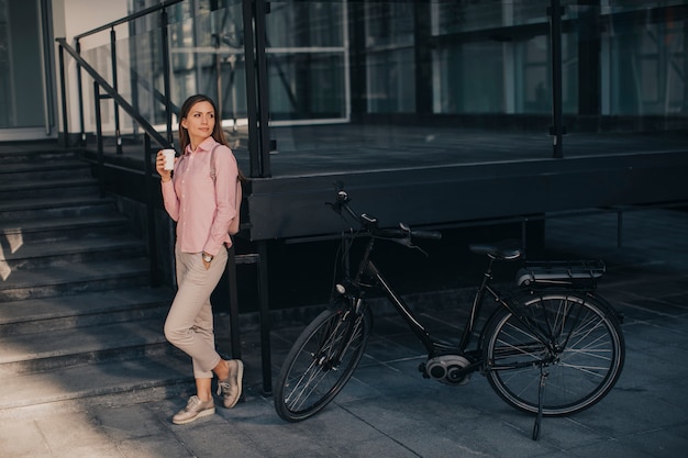 Hermosa joven ciclista bebe café caliente de una taza junto a la bicicleta eléctrica en el entorno urbano