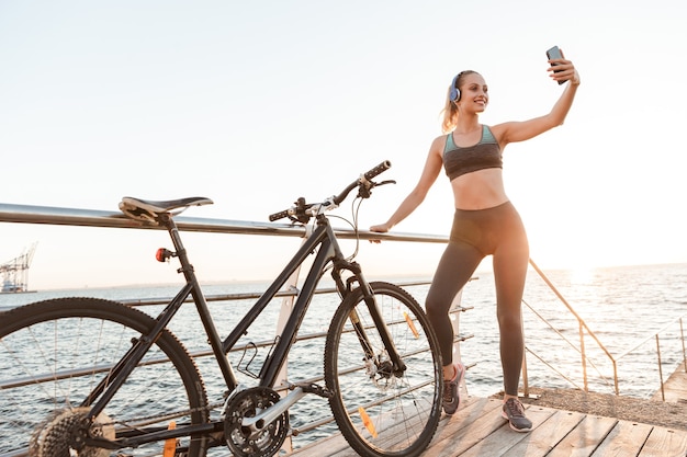 Hermosa joven ciclista de fitness mujer vistiendo ropa deportiva de pie en el muelle con bicicleta, tomando un selfie