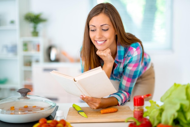 Hermosa joven cocinando comida saludable en la cocina doméstica. Ella está leyendo recetas del libro de cocina.