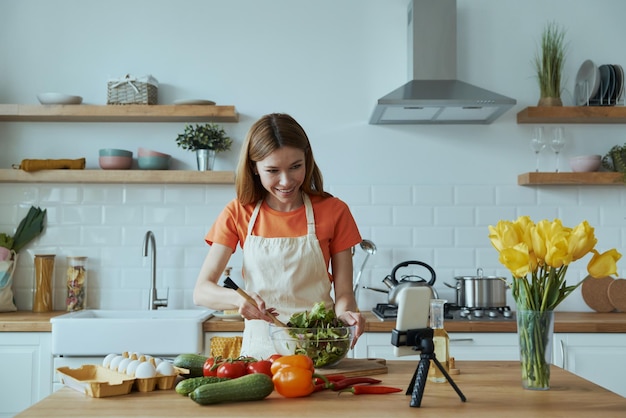 Hermosa joven cocinando y vlogueando mientras está de pie en la cocina doméstica