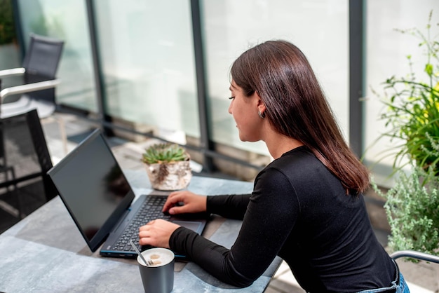 Hermosa joven con una computadora portátil, sentada en la mesa sonriendo y trabajando en una computadora portátil