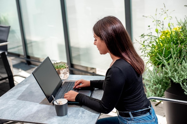 Hermosa joven con una computadora portátil, sentada en la mesa sonriendo y trabajando en una computadora portátil