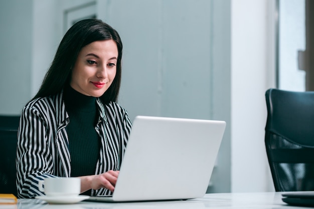 Hermosa joven confiada sonriendo y trabajando en una computadora portátil moderna