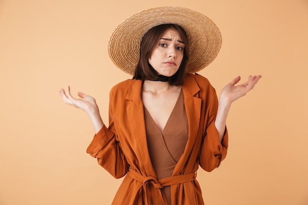 Hermosa joven confundida con sombrero de paja que se encuentran aisladas sobre pared beige, encogiéndose de hombros