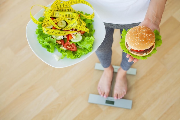 Hermosa joven decide comer hamburguesa o ensalada fresca en la cocina